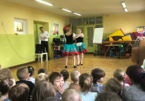 Tancerki tańczą, dzieci siedzą i oglądają.