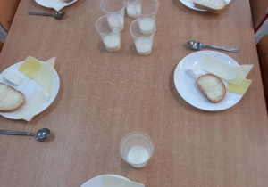 Na stole stoją talerzyki z chlebem i masłem oraz serami, w kubkach stoi kefir.
