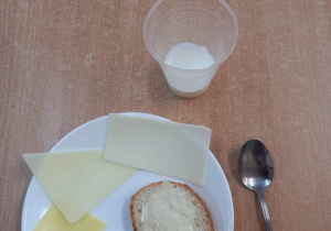 Na stole stoi talerzyk z chlebem i masłem oraz serami, w kubku stoi kefir.