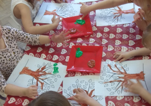 Dzieci malują farbami kontur drzewa i liście używając swoich paluszków.
