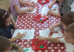 Dzieci malują farbami kontur drzewa i liście używając swoich paluszków.