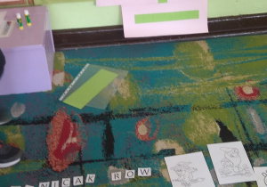 Na dywanie leżą obrazki z postaciami z bajek oraz wycięte kwadraciki z literami. Na tablicy wisi krzyżówka.