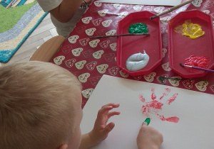 Chłopiec maluje na kartonie zieloną farbą łodygę kwiatu.