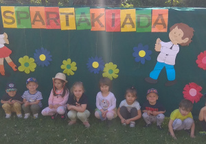 Dzieci z gr I kucają, w tle dekoracja z napisem Spartakiada.