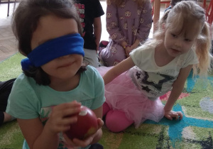 Dziewczynka ma zawiązane niebieską chustą oczy. W rękach trzyma jabłko.