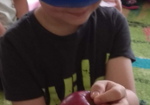 Chłopiec ma zawiązane niebieską chustą oczy. W rękach trzyma jabłko.