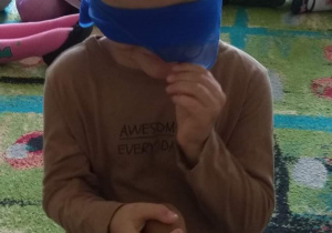 Chłopiec ma zawiązane niebieską chustą oczy. W rękach trzyma kiwi.