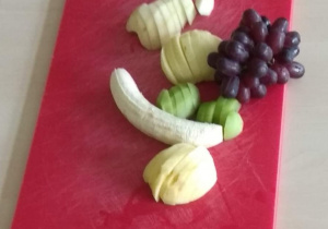 Na desce do krojenia leżą obrane owoce: jabłko, gruszka, kiwi, winogrona, banan.