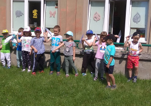 Dzieci stoją wzdłuż murku w ogrodzie przedszkolnym i przygotowują swoje papierowe samoloty do startu:)
