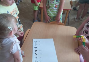 Dzieci stoją wokół stołu, na którym jest karton z ułożonymi podpisami do obrazków.