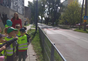 Dzieci stoją za balustradą i wskazują na widoczne po drugiej stronie ulicy znaki - uwaga dzieci, przejście dla pieszych. Pani opowiada o znaczeniu tych znaków.