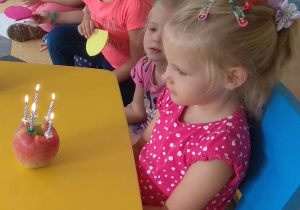 Jubilatka siedzi przy urodzinowym stoliku, na którym ustawiony jest tort - jabłuszko z zapalonymi 5 świeczkami.