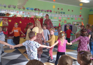 Dzieci tańczą i śpiewają na tle jesiennej dekoracji, przed kolegami i koleżankami z innej grupy.