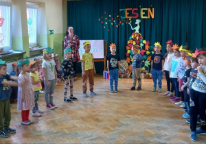 Dzieci z grupy zielonej stoją w półkolu i śpiewają jesienną piosenkę.