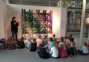 Dzieci siedzą i słuchają opowieści pani o wystawie przypominającej gałęzie.