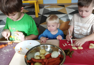 Dzieci kroją warzywa. W środku stoi młodszy brat jednego z przedszkolaków i chce cos podjeść:)
