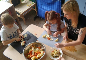 Dzieci kroją warzywa, rodzic przenosi pokrojone warzywa do miski.