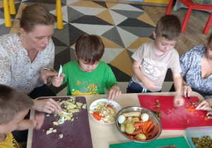 Dzieci wraz z rodzicami i nauczycielem kroją warzywa na sałatkę.