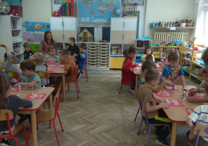 Dzieci siedzą przy stolikach i wyklejają godło Polski białymi piórkami.