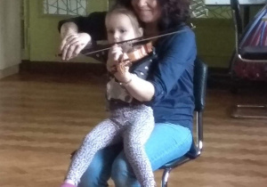 Dziewczynka siedzi na kolanach skrzypaczki i razem z nią gra znaną dzieciom melodię.