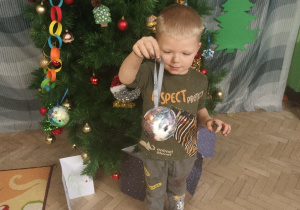Chłopiec stoi przy choince ze swoja bombką ze styropianu.