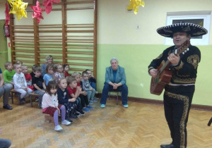 Artysta gra na gitarze, dzieci śpiewają refren znanej piosenki "La cucaracha".