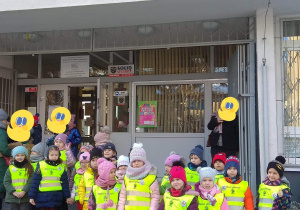 Dzieci w kamizelkach odblaskowych z logo przedszkola stoją przed Szkołą Muzyczną.