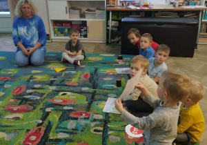 Dzieci siedzą w kole z panią i oglądaja swoje prace.