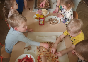 Dzieci nakładają żółty ser na pizzę.
