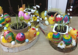 Świąteczny stroik na stół. Na drewnianym plastrze ułożone są: pojemniczek z wysianym owsem, kurczaczek zrobiony z jajka po kinder niespodziance, dwie pisanki malowane przez dzieci i ozdoby.