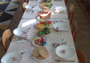Na stołach położony jest biały obrus, a na nim ustawiono talerze, sztućce i świąteczne przysmaki.