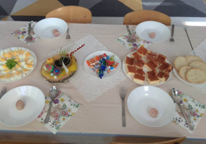Na stołach położony jest biały obrus, a na nim ustawiono talerze, sztućce i świąteczne przysmaki.