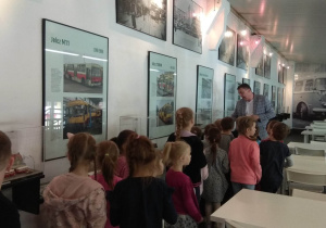 Przewodnik oprowadza dzieci po Muzeum pokazując im zdjęcia dawnych pojazdów komunikacji miejskiej.