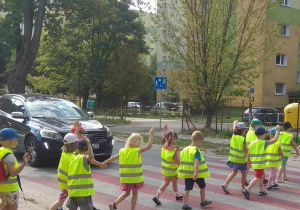 Dzieci z uniesioną ręką przechodzą na pasach na drugą stronę ulicy.
