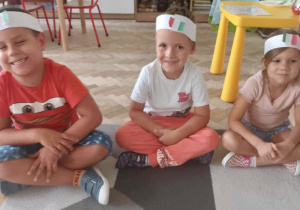Dzieci siedzą na dywanie z opaskami z flagą Włoch na głowie.