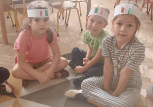 Dzieci siedzą na dywanie w opaskach z flagą Włoch na głowie.