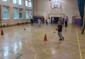 Dzieci rywalizują w dwóch drużynach w zabawie z piłką i pachołkiem.