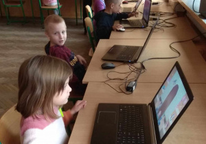Dzieci siedzą przy stanowiskach komputerowych.