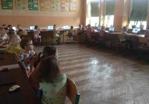 Dzieci siedzą przy stanowiskach komputerowych i z uwagą słuchają Pani Ewy.