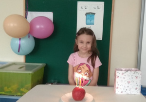 Dziewczynka siedzi na chonorowym miejscu. Na stoliku stoi tort z jabłuszka.