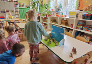 Dzieci słuchają bajki o trzech świnkach, kolejno podchodzą i dokładają rekwizyty na scenę.