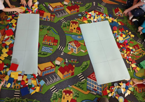 Na dywanie leżą dwa duże kartony podzielone na kilka pól oraz kolorowe plastikowe figury geometryczne.