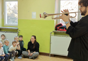Dzieci słuchają dźwięków wygrywanych na trąbce.