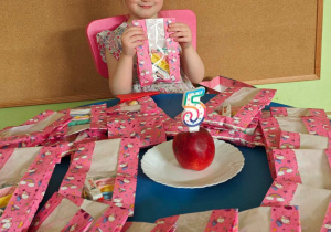 Jubilatka siedzi przy stoliku zastawionym upominkami dla dzieci.